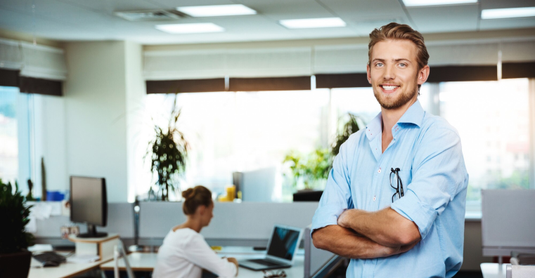 Young successful businessman smiling, posing with crossed arms, over office